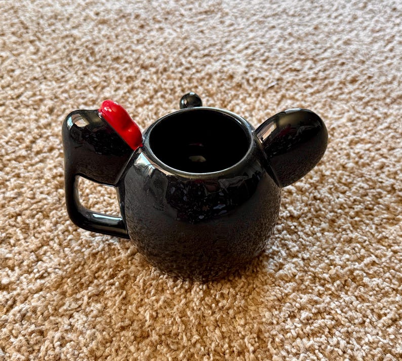 May include: Black ceramic mug shaped like a character's head, featuring large ears and a red bow. The mug has a handle and a glossy finish, perfect for hot beverages.