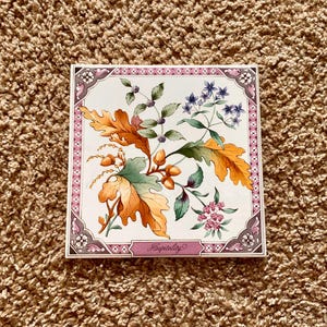 May include: A white ceramic tile with a pink border and a floral design. The tile features a variety of flowers and leaves in shades of yellow, green, blue, and pink. The tile also has the word "Hospitality" written in pink lettering.