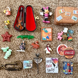 May include: A collection of miniature items including a violin in a case, a Minnie Mouse figure, a brown suitcase, a red star, a green lizard, a framed abstract print, a Coca-Cola button, a My Fair Lady card, a metal spur, and various small figurines.