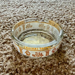 May include: Clear glass ashtray with a gold rim and a white band featuring the text "CAESARS TAHOE" and gold illustrations of figures. The ashtray is circular and sits on a brown carpet.