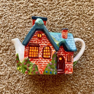 May include: A red brick house shaped teapot with blue and green accents. The teapot has a white spout and handle. The house has two windows, a door, and a chimney.