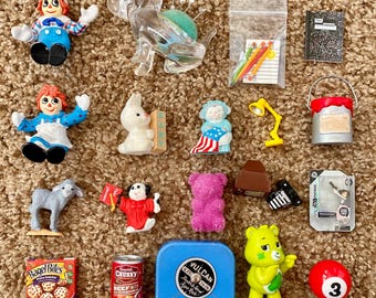 May include: A collection of miniature toys and collectibles, including Raggedy Ann dolls, a clear plastic sewing machine, a notepad, a paint can, a teddy bear, a pool ball, and a can of Chunky Beef with vegetables.