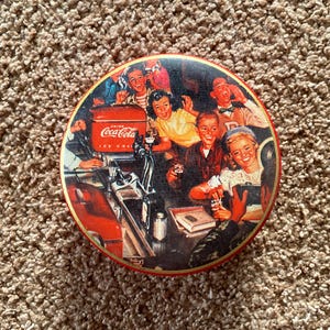 Puede incluir: Una lata vintage de Coca-Cola con una ilustración colorida de un grupo de personas disfrutando de una bebida en una fuente de soda. La lata presenta el logotipo de Coca-Cola y el texto "Drink Coca-Cola Ice Cold".