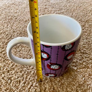 Puede incluir: Taza de cer&aacute;mica blanca con un fondo morado con un patr&oacute;n repetido de la cara de un personaje. La taza mide aproximadamente 10 cm de alto y tiene un asa blanca.