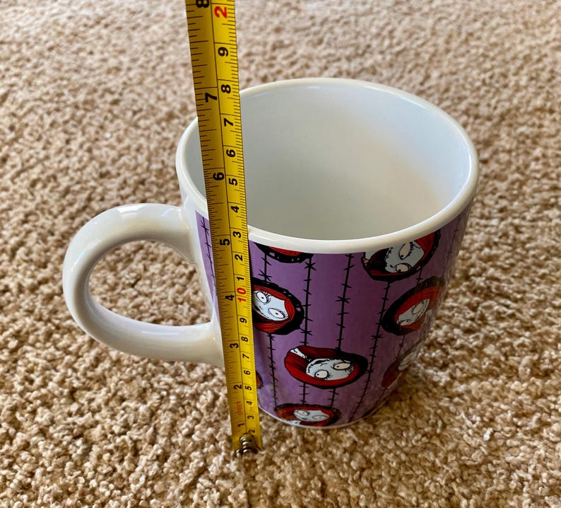 Puede incluir: Taza de cer&aacute;mica blanca con un fondo morado con un patr&oacute;n repetido de la cara de un personaje. La taza mide aproximadamente 10 cm de alto y tiene un asa blanca.