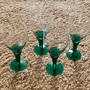 May include: Four clear wine glasses with emerald green stems and bases. The glasses are empty and arranged on a textured, light brown surface. The green bases add a pop of color to the elegant glassware.