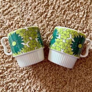 Könnte beinhalten: Zwei weiße Keramikbecher mit einem grün-gelben Blumenmuster. Die Becher haben ein gestapeltes Design mit geripptem Boden und gebogenem Griff. Das Blumenmuster zeigt große grüne Gänseblümchen und kleinere gelbe Blüten.