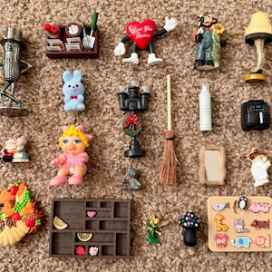 Puede incluir: Una colección de figuras en miniatura y objetos decorativos, que incluyen un personaje en forma de corazón, un conejito, una lámpara, una cornucopia, una escoba de bruja y una mini freidora. Los artículos están dispuestos sobre una alfombra marrón.