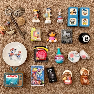 May include: A collection of miniature toys and trinkets including a metal key, Disney figurines, blue blocks with character prints, a Dora the Explorer figure, a black 8-ball, a small plate, a Toy Story DVD case, a Jaws television toy, and various charms.