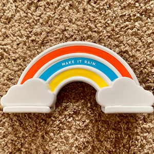 May include: A ceramic rainbow with orange, blue, and yellow bands, white clouds, and the text "MAKE IT RAIN." The rainbow is a decorative item, possibly a shelf sitter or a small sculpture.