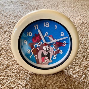 May include: A round, cream-colored alarm clock with a blue face featuring the Tasmanian Devil cartoon character. The clock has white hands and black numerals. The image shows the clock on a brown carpet.
