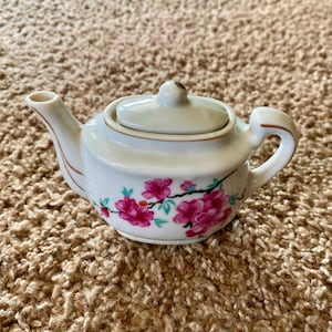 May include: A small, white ceramic teapot with a lid and handle. The teapot is decorated with pink floral designs and gold trim. The spout is curved, and the handle is arched. A charming piece for tea lovers.