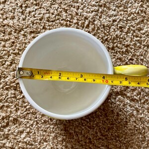 May include: A white ceramic mug with a yellow handle is measured with a tape measure. The mug's diameter is approximately 4 inches. The interior of the mug is white, and the handle is a bright yellow color.