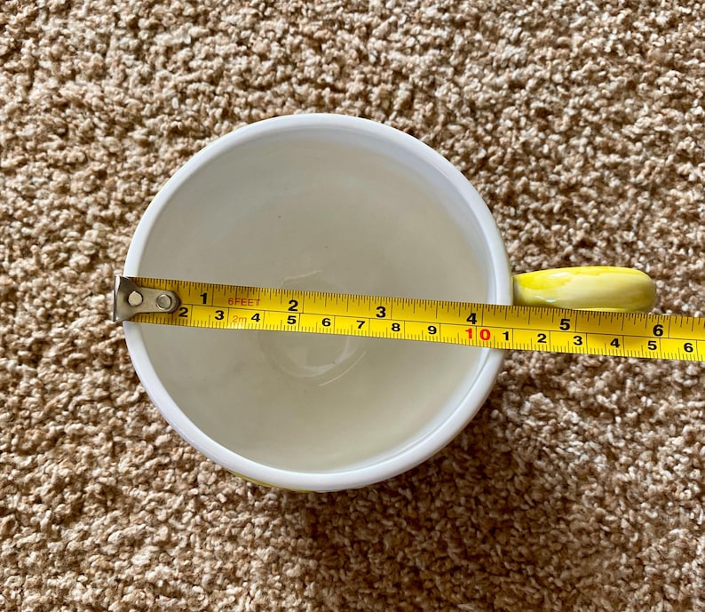 May include: A white ceramic mug with a yellow handle is measured with a tape measure. The mug's diameter is approximately 4 inches. The interior of the mug is white, and the handle is a bright yellow color.