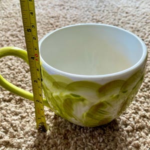 May include: A large, white ceramic mug with a green handle and a decorative green painted design. The mug is approximately 4 inches tall, measured with a yellow tape measure.