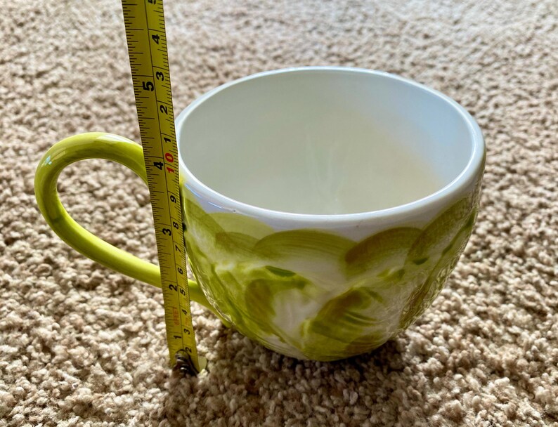 May include: A large, white ceramic mug with a green handle and a decorative green painted design. The mug is approximately 4 inches tall, measured with a yellow tape measure.