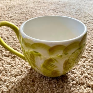 May include: A ceramic coffee mug with a bright yellow handle and a white interior. The exterior is decorated with a pattern of olive green brushstrokes. The mug is designed for hot beverages.