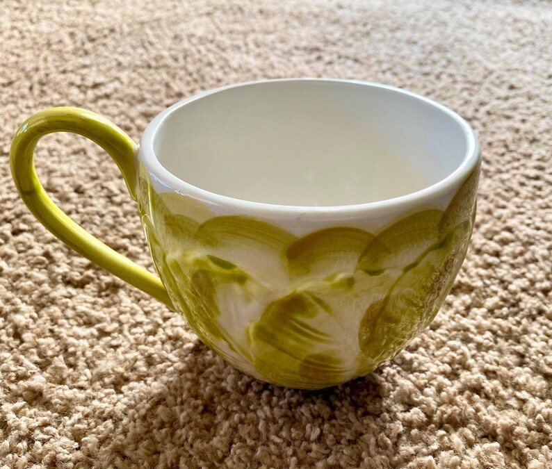 May include: A ceramic coffee mug with a bright yellow handle and a white interior. The exterior is decorated with a pattern of olive green brushstrokes. The mug is designed for hot beverages.