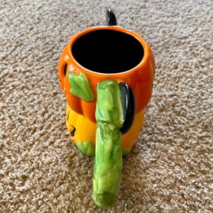 以下が含まれることがあります： カボチャの形をした、ハロウィーンをテーマにしたセラミックマグ。マグはオレンジ色で、緑の葉のアクセントと緑のハンドルが付いています。マグの内側は黒です。マグは茶色のカーペットの上に置かれています。