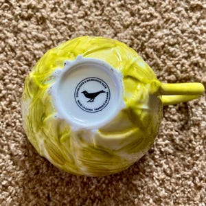 May include: A yellow ceramic teacup with a textured, leafy design. The handle is a solid yellow. The bottom of the cup is white with a black bird logo and the text "Dishwasher & Microwave Safe Made in China Fabriqu&eacute; en Chine."