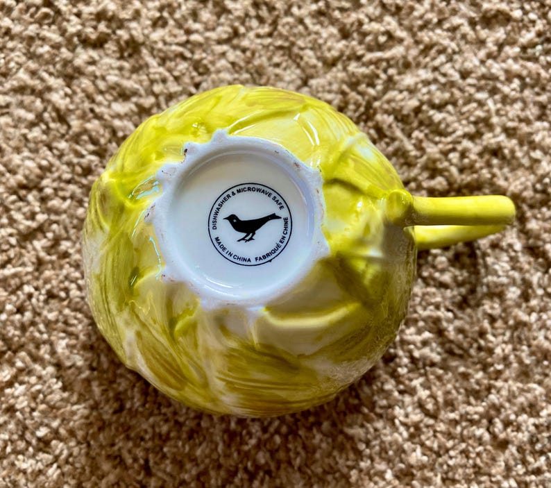 May include: A yellow ceramic teacup with a textured, leafy design. The handle is a solid yellow. The bottom of the cup is white with a black bird logo and the text "Dishwasher & Microwave Safe Made in China Fabriqu&eacute; en Chine."