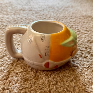 May include: A ceramic mug featuring a design inspired by a character from a popular science fiction series. The mug has a light gray handle and body, with orange and green accents. The mug is sitting on a brown carpet.