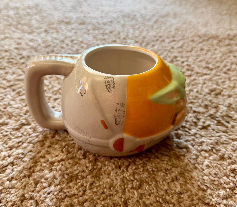 May include: A ceramic mug featuring a design inspired by a character from a popular science fiction series. The mug has a light gray handle and body, with orange and green accents. The mug is sitting on a brown carpet.