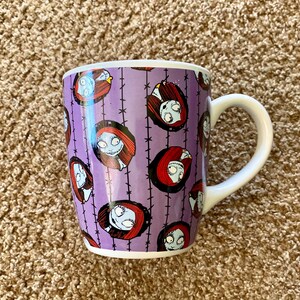 Puede incluir: Una taza de cer&aacute;mica con asa y borde blancos. La taza presenta un fondo morado con un patr&oacute;n repetido de Sally de The Nightmare Before Christmas. El dise&ntilde;o incluye la cara y el pelo rojo de Sally.