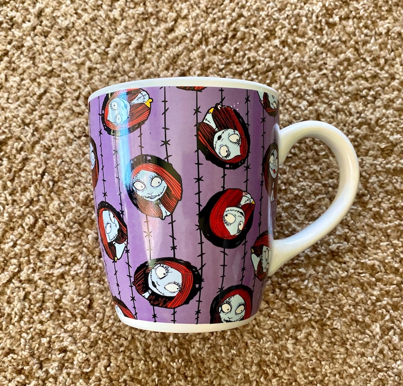 Puede incluir: Una taza de cer&aacute;mica con asa y borde blancos. La taza presenta un fondo morado con un patr&oacute;n repetido de Sally de The Nightmare Before Christmas. El dise&ntilde;o incluye la cara y el pelo rojo de Sally.