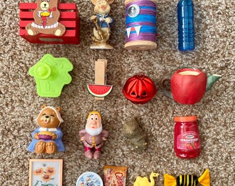 May include: A collection of miniature figurines and objects, including a red bear in a wooden crate, a pig figurine, a spool of thread, a water bottle, a green object, a watermelon clip, a red bell, an apple teapot, a bear figurine, a dwarf figurine, a jar of strawberry jam, a picture of sunflowers, a container of Cool Whip, a caramel wrapper, a camel figurine, and a candy.