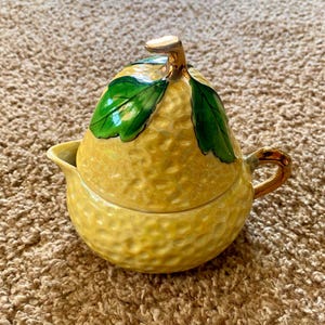 Puede incluir: Un juego de azucarero y crema de cerámica amarilla con forma de pera. La crema tiene un pico y un asa, y el azucarero tiene una tapa con hojas verdes. El juego tiene una superficie texturizada.