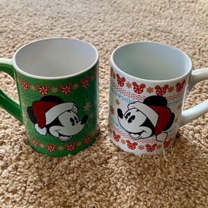 Könnte beinhalten: Zwei Keramikbecher mit Mickey Mouse mit Weihnachtsmütze. Ein Becher ist grün mit roten und weißen Akzenten, der andere ist weiß mit roten Akzenten. Beide Becher haben einen weißen Innenraum und einen gebogenen Griff.