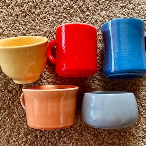 Puede incluir: Cinco tazas de cerámica coloridas en tonos amarillo, rojo, azul, naranja y gris. Las tazas varían en forma, algunas con asas y otras sin ellas. Las tazas están dispuestas sobre una superficie marrón texturizada.