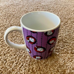 Puede incluir: Una taza de cer&aacute;mica blanca con un exterior morado que presenta un patr&oacute;n repetido de la cara de un personaje de la pel&iacute;cula "Pesadilla antes de Navidad". La taza tiene un asa e interior blancos.