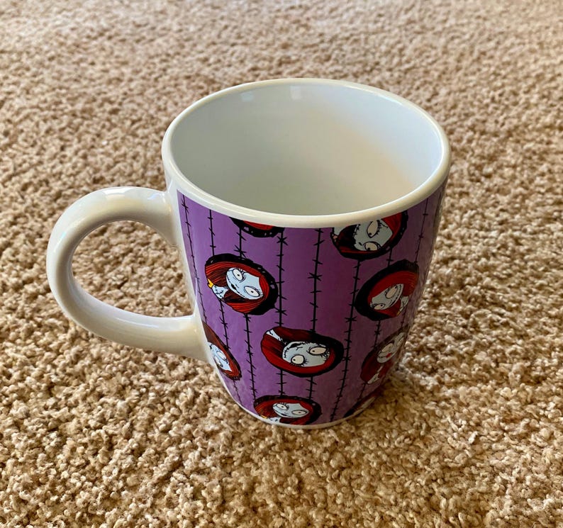 Puede incluir: Una taza de cer&aacute;mica blanca con un exterior morado que presenta un patr&oacute;n repetido de la cara de un personaje de la pel&iacute;cula "Pesadilla antes de Navidad". La taza tiene un asa e interior blancos.