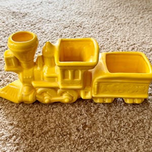 May include: A bright yellow ceramic train-shaped planter featuring a locomotive engine with a smokestack opening and a connected carriage with a rectangular open compartment. The piece has molded wheel details and a smooth, glossy finish throughout.