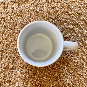 May include: Overhead view of a white ceramic mug with a handle. The interior of the mug is a light blue color. The mug is sitting on a brown, textured surface.