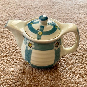 Könnte beinhalten: Eine Keramik-Teekanne mit Deckel, Henkel und Ausguss. Die Teekanne ist mit einem blau-weiß gestreiften Muster bemalt, das mit kleinen floralen Details akzentuiert ist. Der Deckel hat einen blauen Knopf und ein passendes blau-weißes Design.