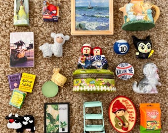 Peut inclure: Une collection de figurines et de bibelots miniatures, dont une poupée, une boîte Spider-Man, une peinture de paysage, une théière, un agneau, une boule de billard, une figurine de méchant, un chien, une vache, une plante miniature, un chariot miniature et un paquet de bonbons Haribo.