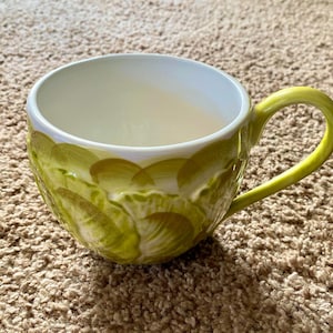 May include: A ceramic mug with a wide, rounded bowl and a curved handle. The mug is primarily light green with a textured pattern of overlapping brushstrokes in shades of green and yellow. The interior is white, and the handle is a bright yellow-green.