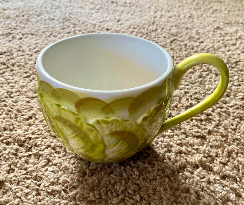 May include: A ceramic mug with a wide, rounded bowl and a curved handle. The mug is primarily light green with a textured pattern of overlapping brushstrokes in shades of green and yellow. The interior is white, and the handle is a bright yellow-green.