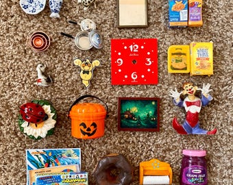 May include: A collection of miniature decorative items, including a red clock face, a clown figurine, a miniature painting, and a jar of grape jelly. Other items include a ladybug on a flower, a Halloween bucket, and a vintage Aquaman game.