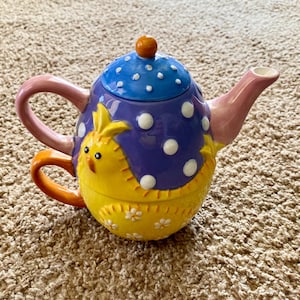 Puede incluir: Una tetera de cerámica caprichosa con un diseño de pollito. La tapa es azul con lunares blancos y un pequeño botón naranja. El cuerpo es amarillo con un pollito, y el asa y el pico son rosas y naranjas.