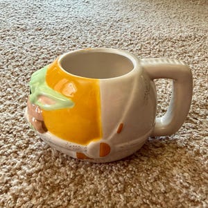 May include: A ceramic mug featuring a design inspired by a popular character. The mug is primarily white with a yellow and green design on the front, and a large handle. The mug is designed to resemble a character's head.