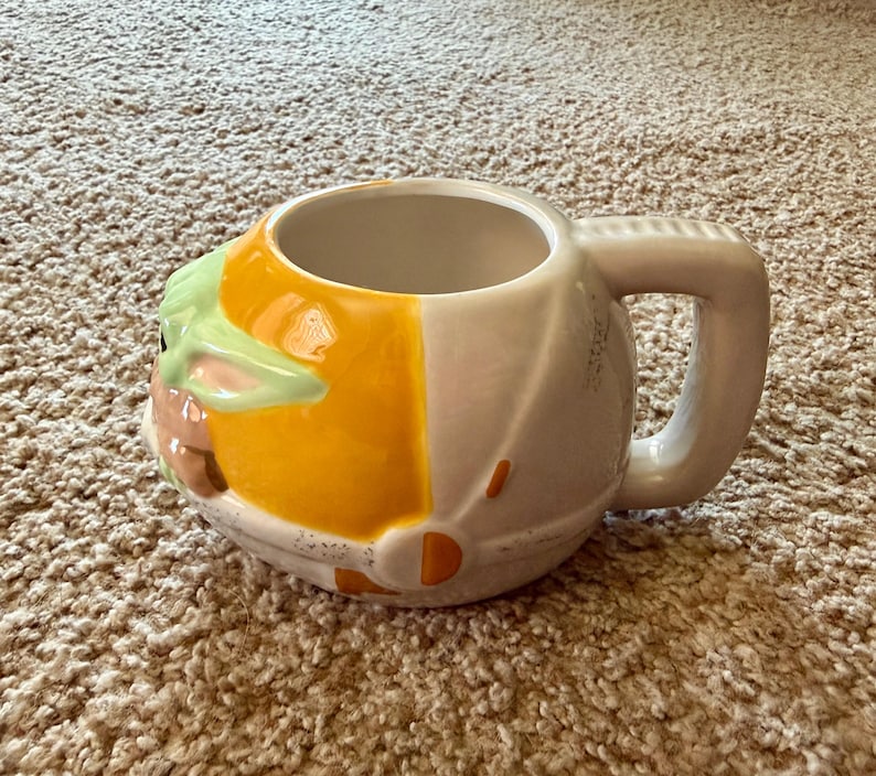 May include: A ceramic mug featuring a design inspired by a popular character. The mug is primarily white with a yellow and green design on the front, and a large handle. The mug is designed to resemble a character's head.