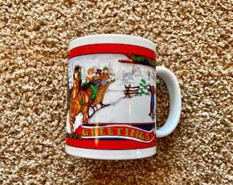 May include: White ceramic mug with a winter scene featuring a horse-drawn sleigh and the word "Greetings" in red and gold lettering.