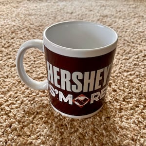 May include: White ceramic mug with a brown background and the text "Hershey's S'mores" in white letters.