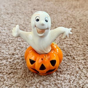 May include: A ceramic Halloween salt and pepper shaker set. The set features a smiling white ghost figure atop an orange jack-o'-lantern. The ghost has black eyes and a smiling mouth. The jack-o'-lantern has carved-out eyes and a mouth.