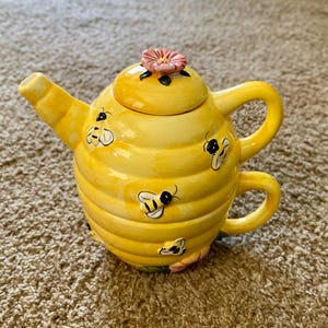 Op de afbeelding: Een gele keramische theepot en kopjes set, ontworpen om op een bijenkorf te lijken. De theepot heeft een deksel met een roze bloem en is versierd met cartoon bijen. De set is ontworpen voor één persoon.