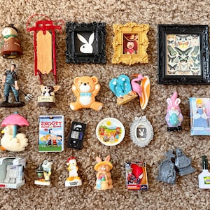 May include: A collection of miniature figurines and decorative items including a red sled, framed art, a teddy bear, a Snoopy VHS case, a gnome, a duck with an umbrella, a rabbit, a Spider-Man box, and various other small plastic collectibles.
